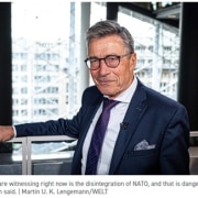 Man in a navy suit and glasses speaks in a bright studio; caption quotes Rasmussen: NATO disintegration is dangerous.